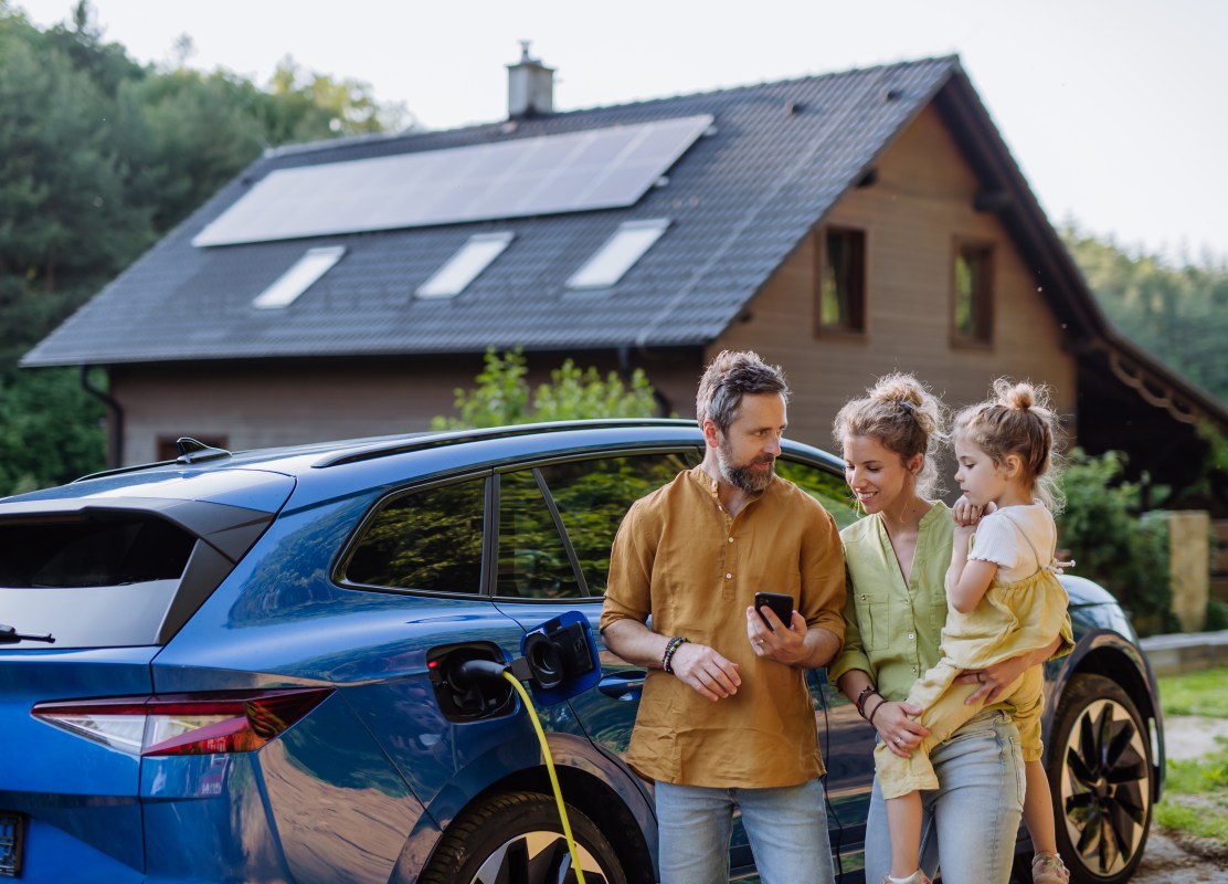 Family with little girl standing in front of their house with solar panels on the roof, having electric car.